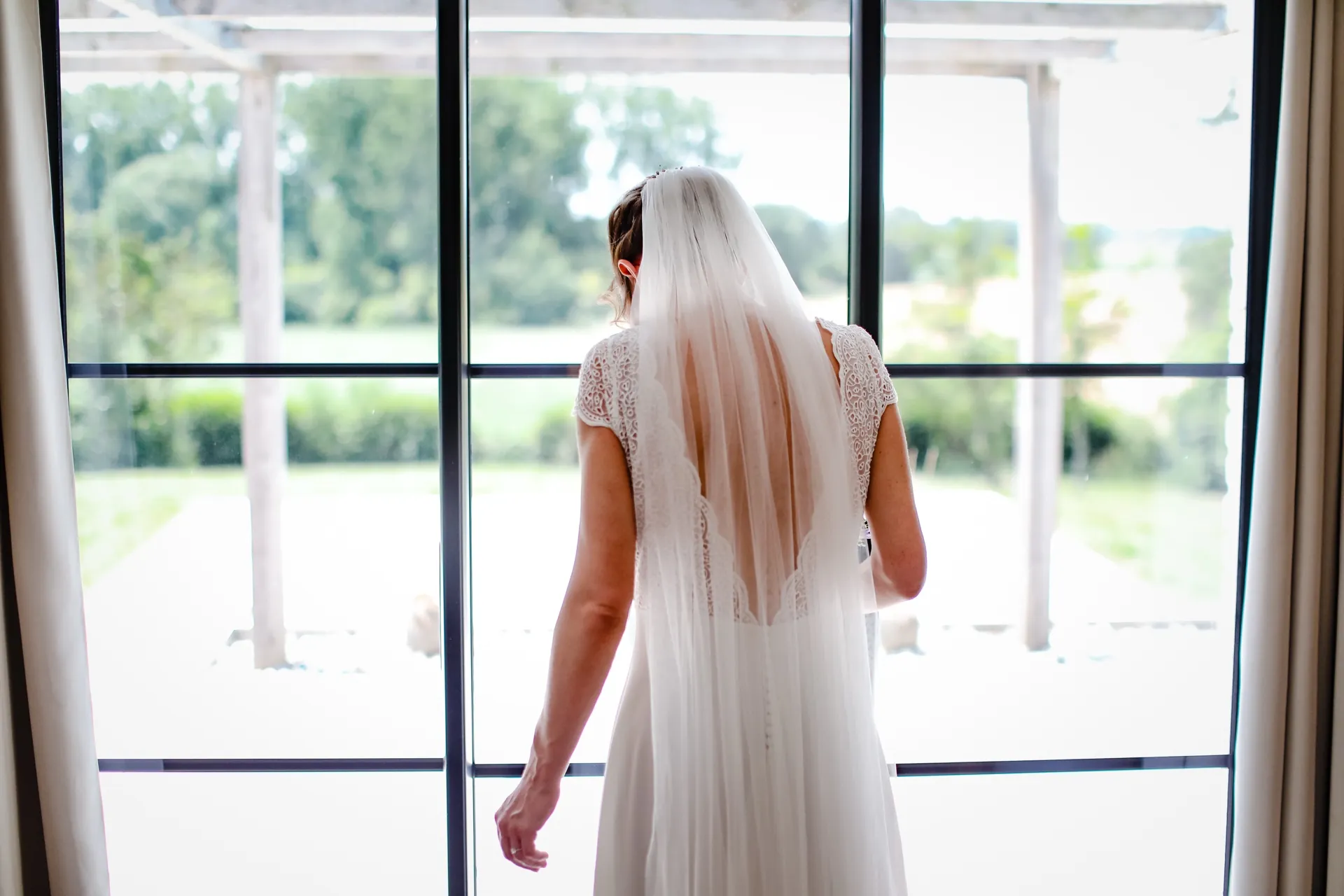 Aurélie de dos devant la porte-fenêtre, voile long et dos en V de la robe KATE