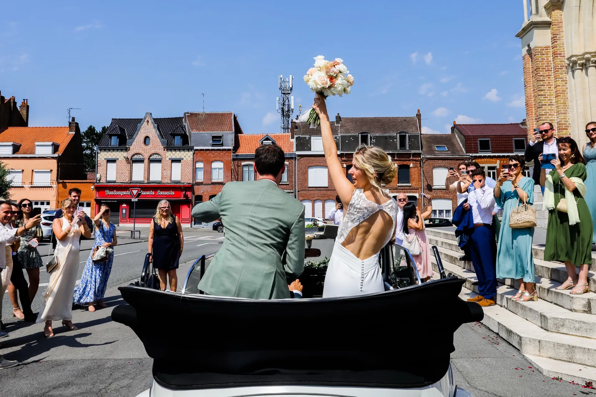 Lola, bouquet levé, salue les invités depuis la voiture des mariés à la sortie de l'église : dos échancré en tulle brodé
