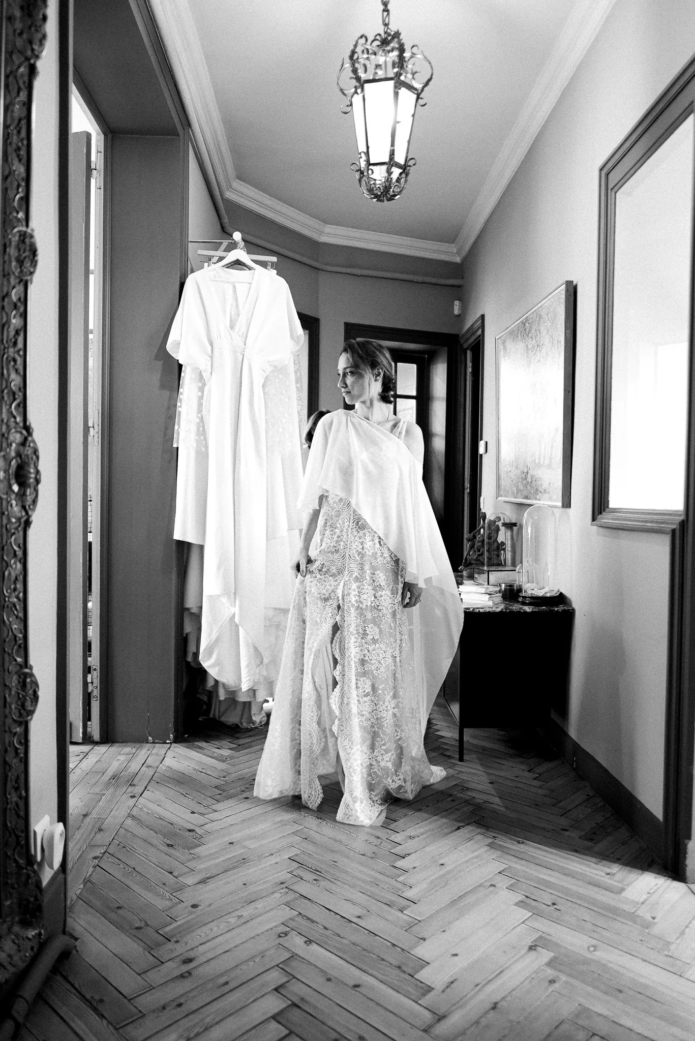 Marie dans le couloir du showroom, la robe en dentelle sous une cape légère, en noir et blanc